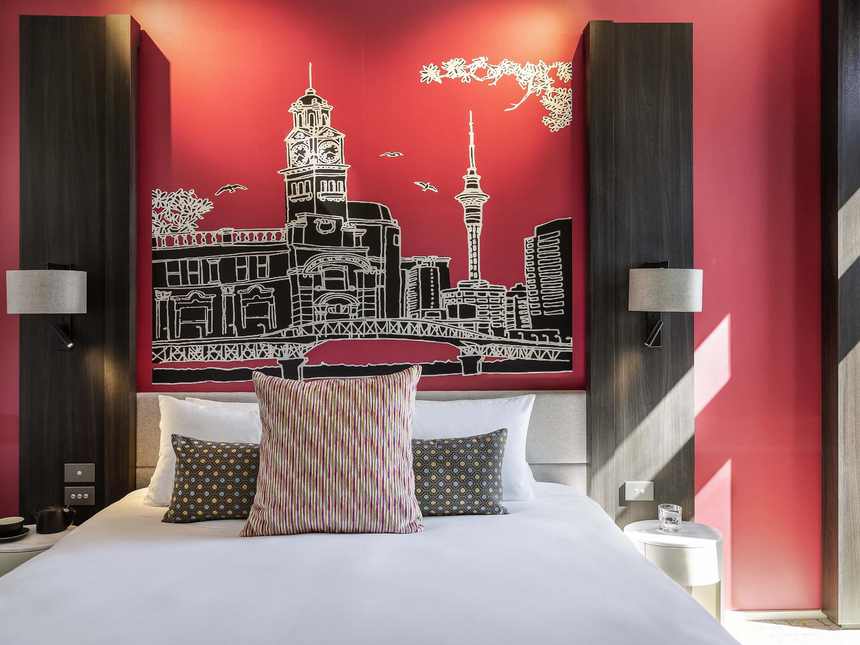 A hotel room with a bed, pillows, a nightstand, a wall art of a cityscape, and two wall-mounted lamps.