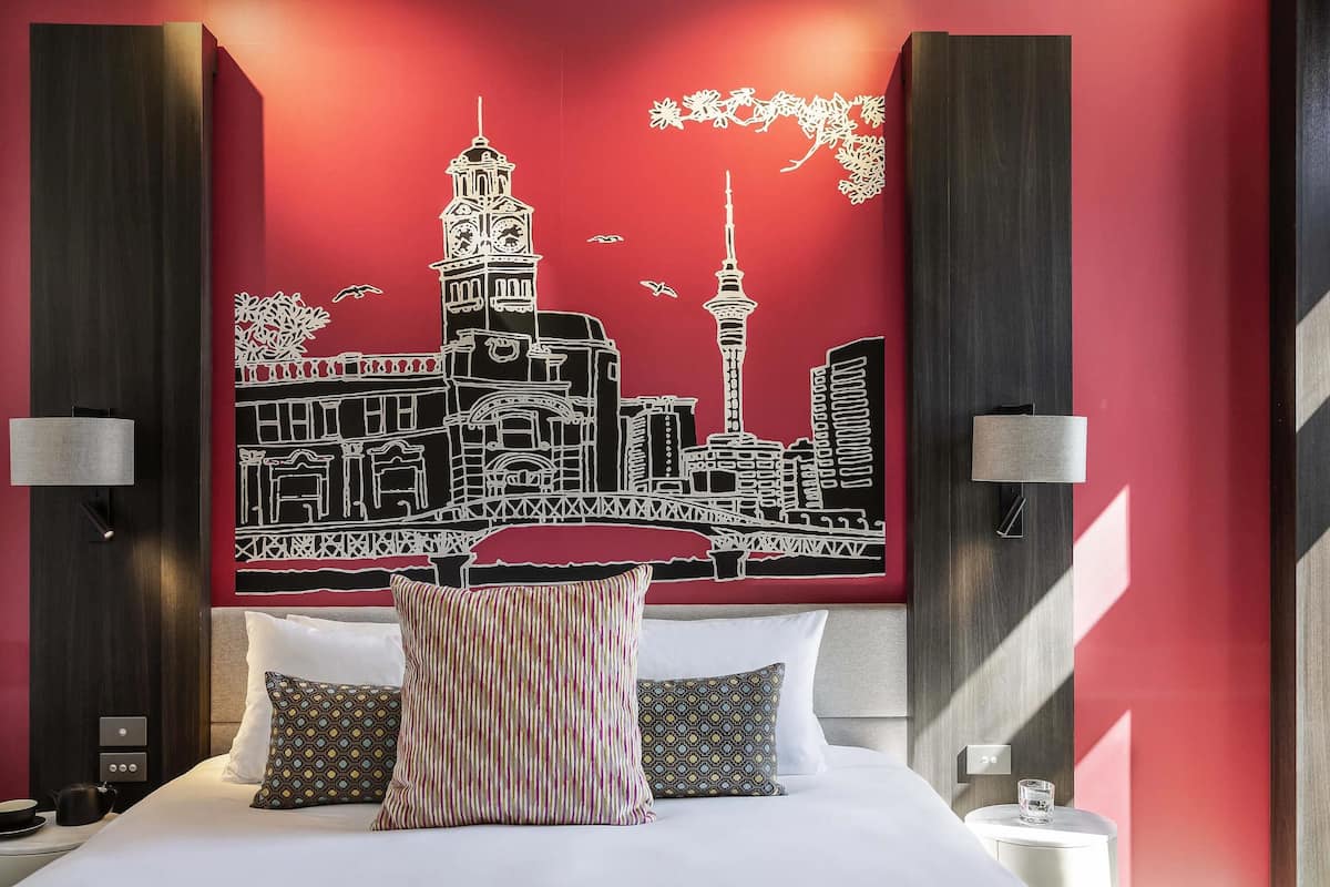 A hotel room with a bed, pillows, a nightstand, a wall art of a cityscape, and two wall-mounted lamps.