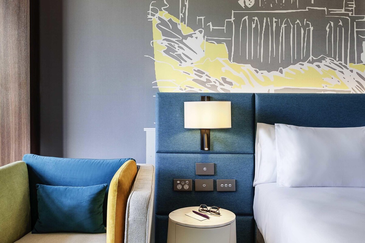 A hotel room with a bed, a nightstand with a lamp, a chair with cushions, and a wall with a cityscape mural.