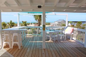 Una terraza de madera con muebles de exterior blancos, incluyendo una mesa y sillas, con vista a una playa con palmeras y un edificio al fondo.