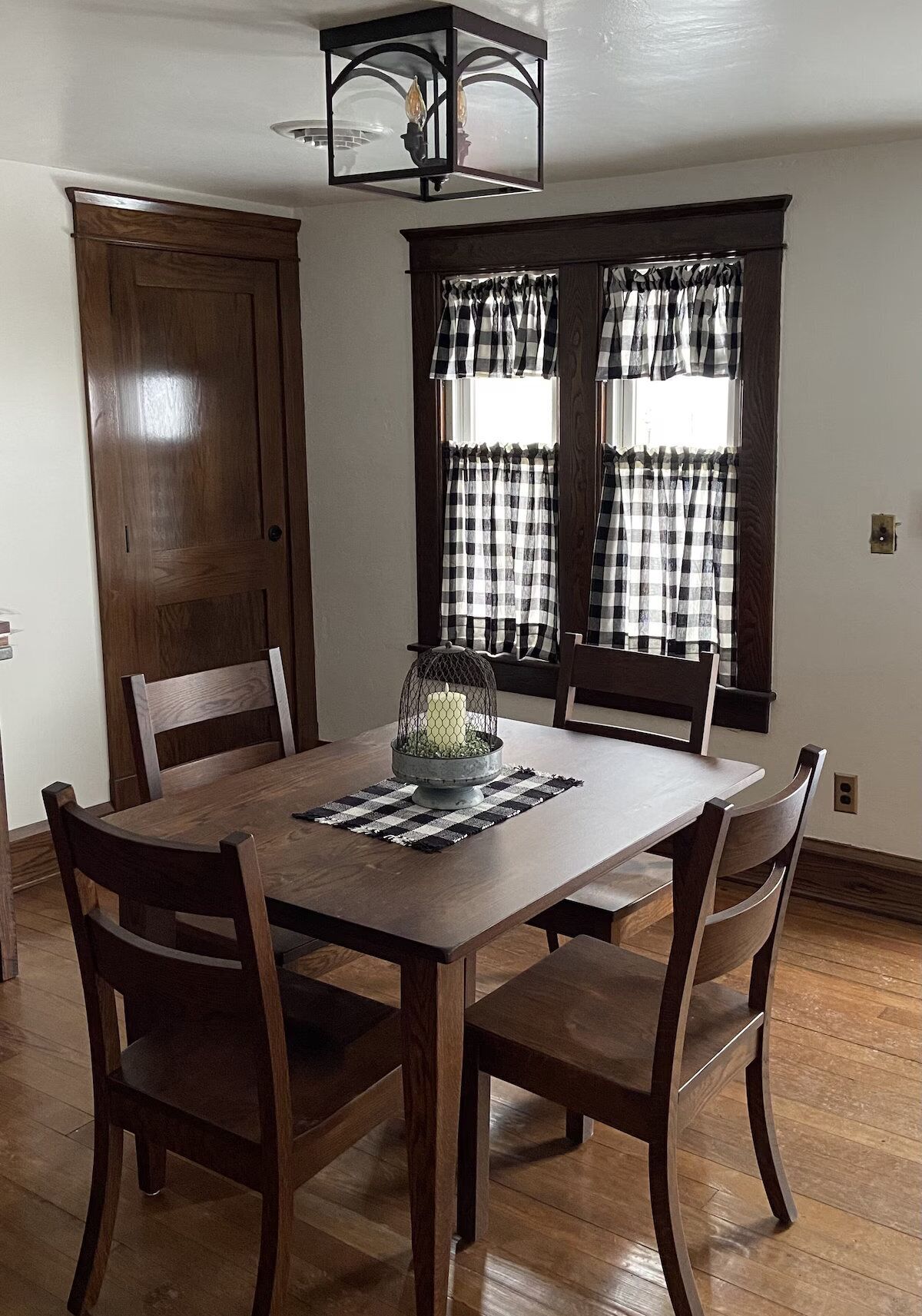 Comedor con mesa de madera, cuatro sillas, candelabro y dos ventanas con cortinas a cuadros.