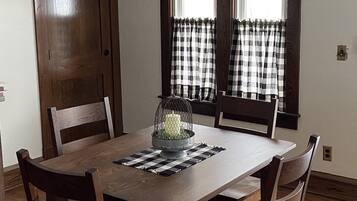 Comedor con mesa de madera, cuatro sillas, candelabro y dos ventanas con cortinas a cuadros.