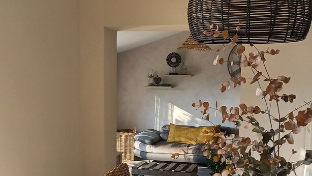 A dining area with a round table, wicker chairs, a woven ceiling light fixture, and a vase with dried flowers.