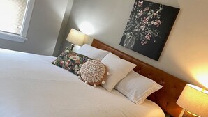 En snyggt bäddad säng med vita sängkläder, en kudde med blommönster och en sänggavel med en tavla ovanför. Det finns två sänglampor och en mönstrad matta på golvet.