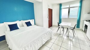 Een hotelkamer met een bed, een eettafel met stoelen, een keukenhoek en uitzicht op zee door een raam.