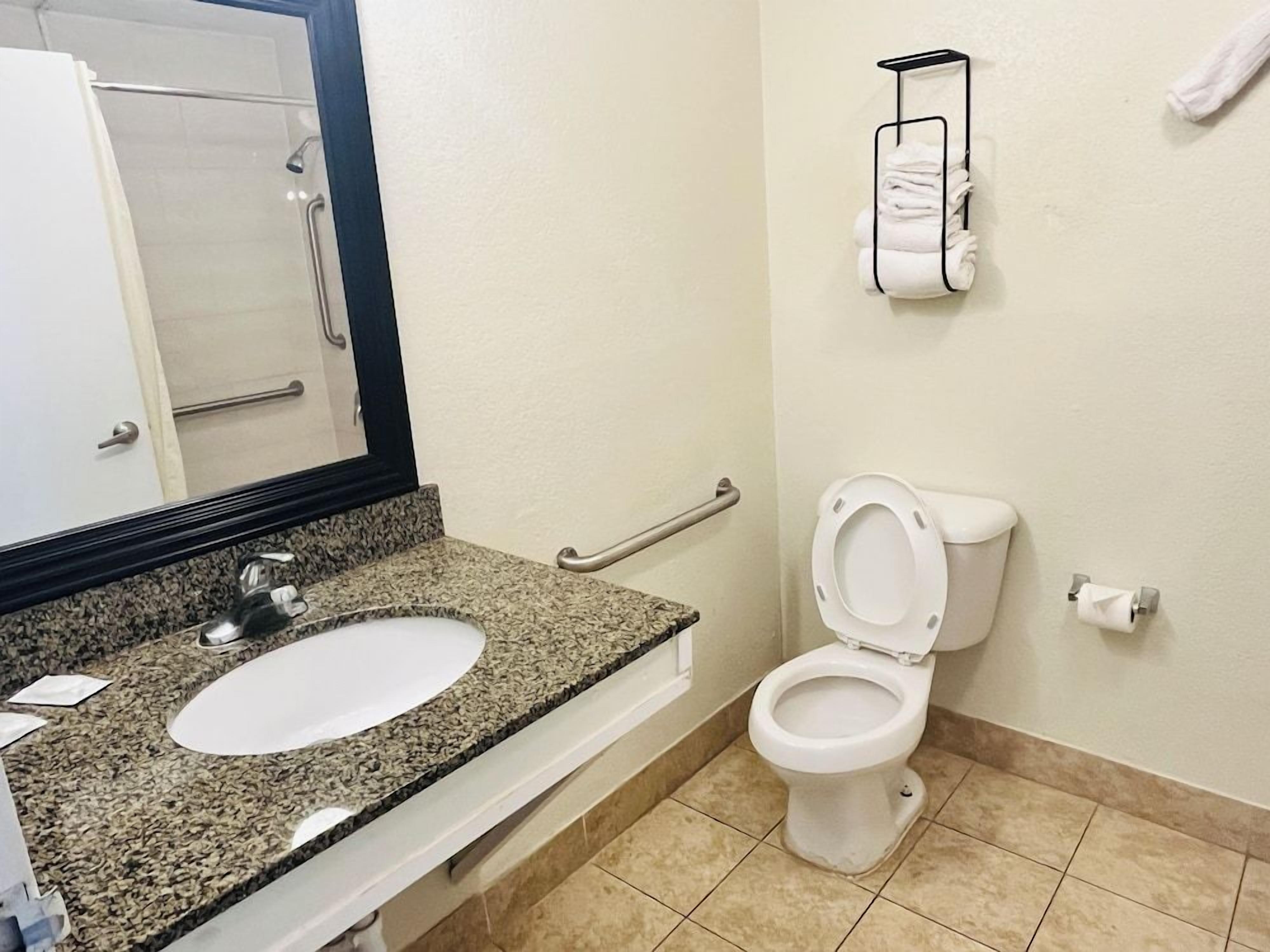 A bathroom with a granite countertop sink, a white toilet, a towel rack, and a handrail.