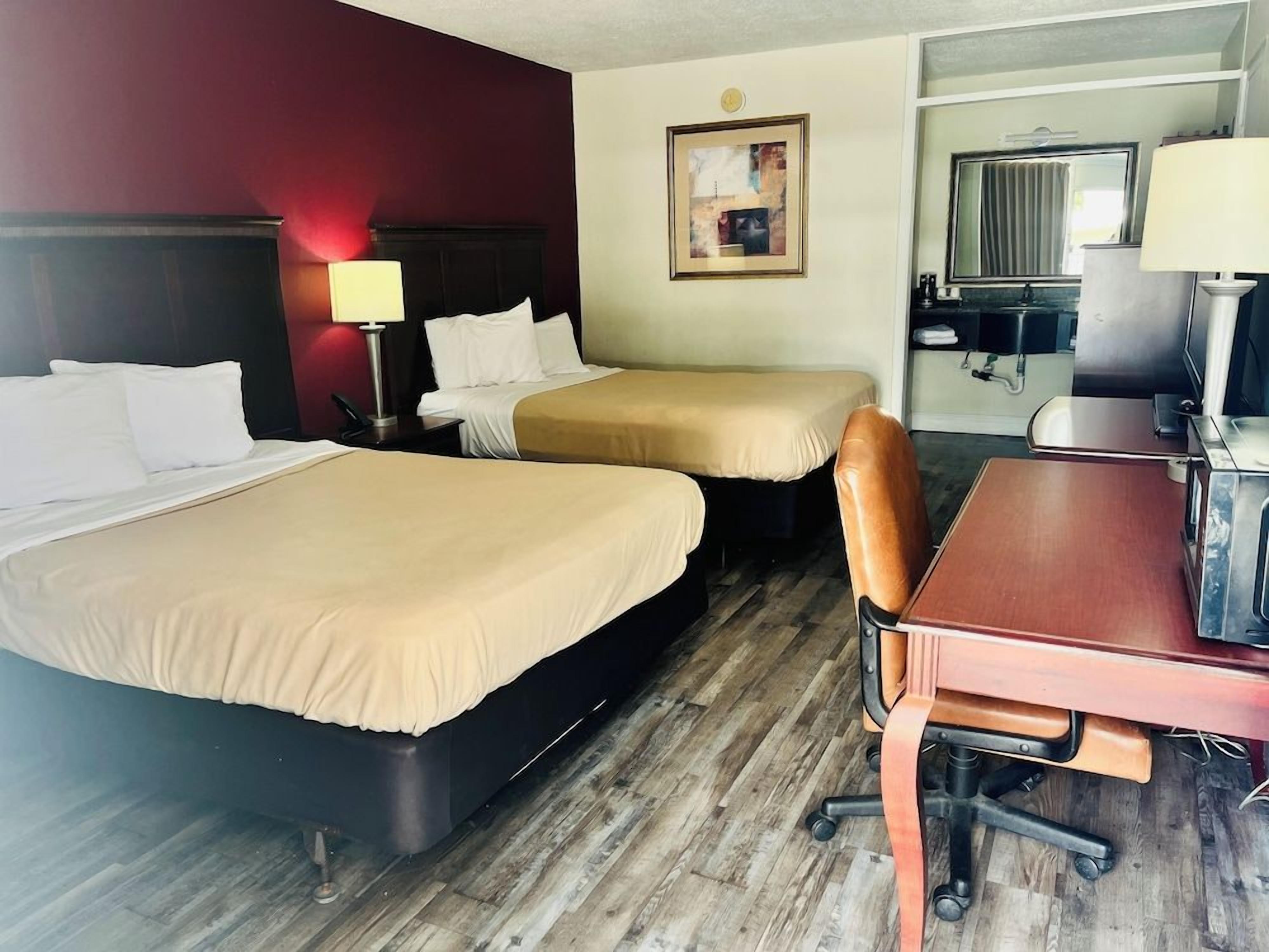 A hotel room with two beds, a desk, a chair, a lamp, a painting, and a mirror.