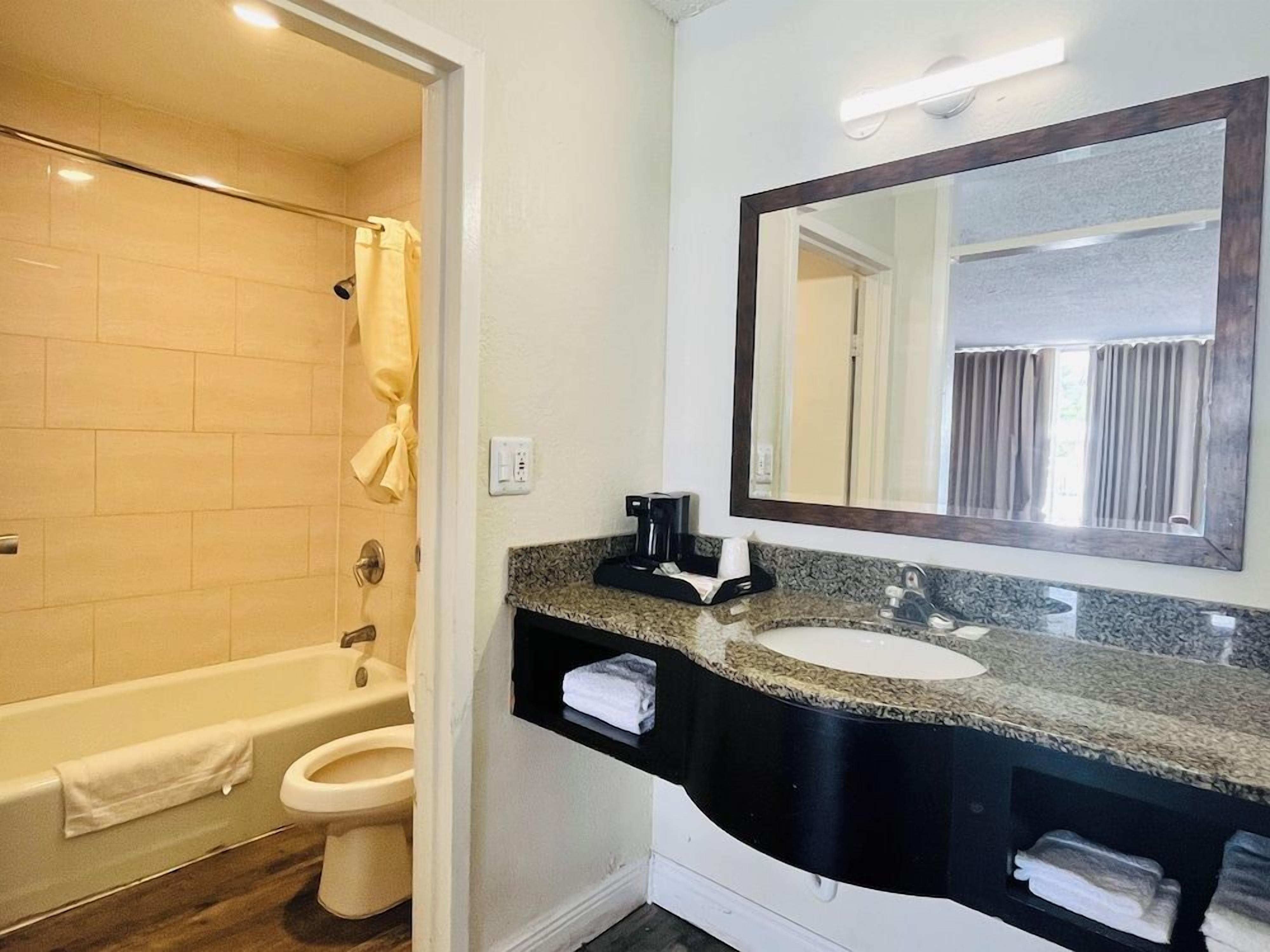A bathroom with a bathtub, toilet, and sink. There is a towel rack with towels, a mirror, and a coffee maker on the counter.