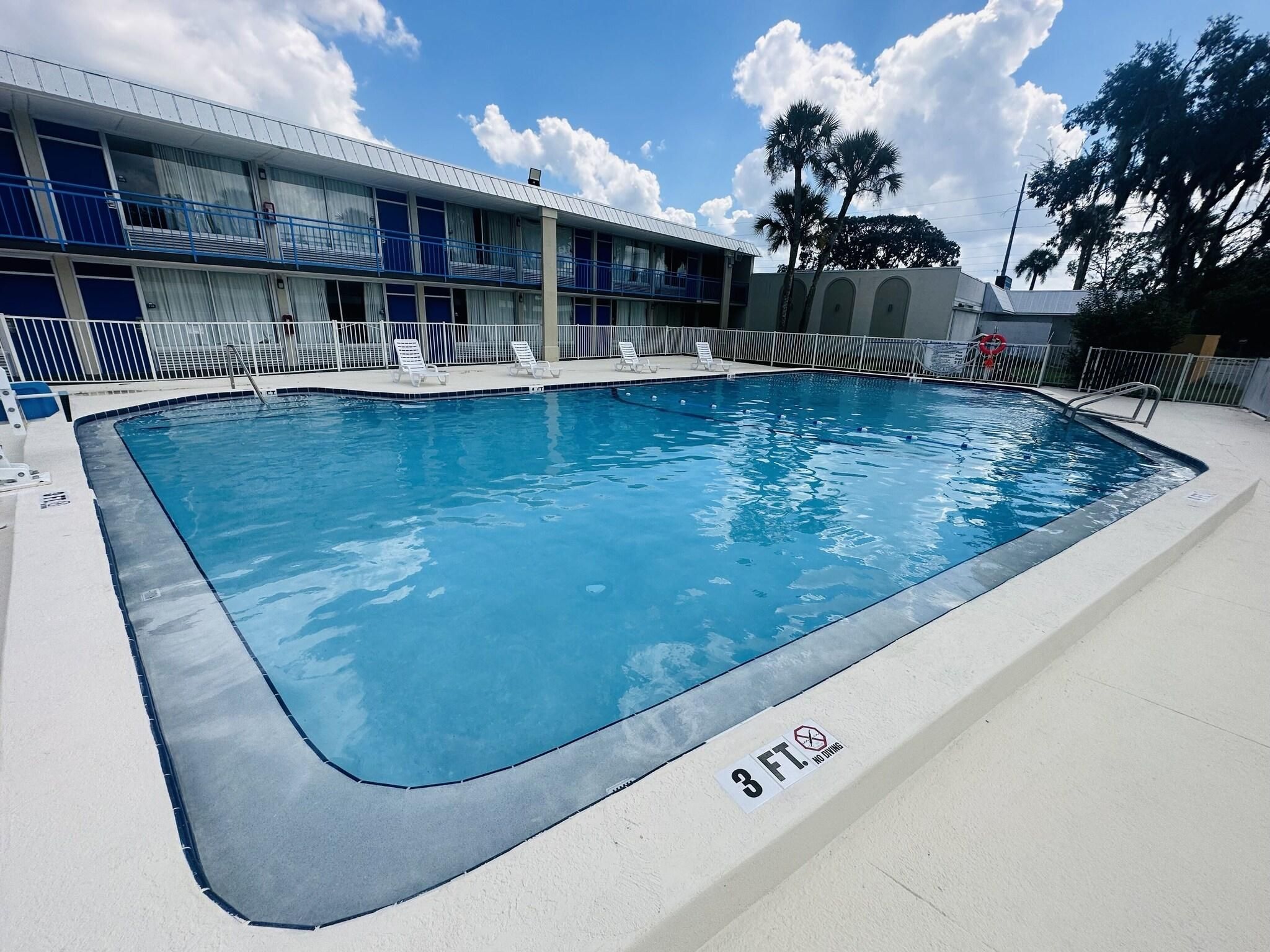 Uma piscina retangular com um aviso de 3 FT, proibido mergulhar, rodeada por um deck branco e um edifício com varandas azuis.