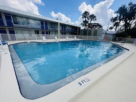 Una piscina rectangular con un letrero de 3 pies que indica 'no saltos', rodeada por un patio blanco y un edificio con balcones azules.