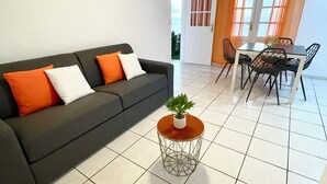 Un salon moderne avec un canapé gris, des coussins orange et blancs, une table basse ronde agrémentée d’une plante, et un coin repas avec une table et des chaises.