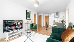 Ein modernes Wohnzimmer mit Flachbildfernseher, Glastisch, grünem Sofa und einem Essbereich mit weißem Tisch und Stühlen.