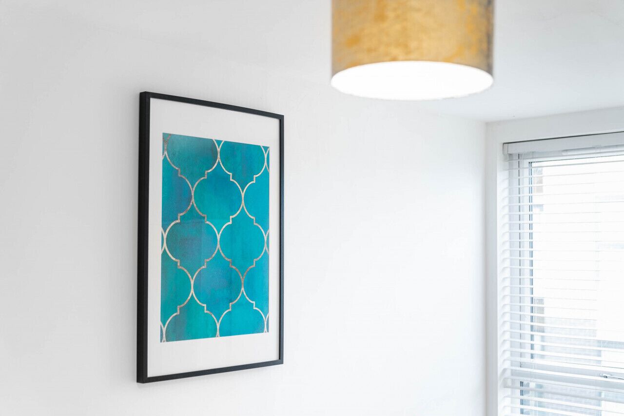 Una obra de arte abstracto enmarcada sobre una pared blanca, una ventana con persianas y una lámpara colgante.