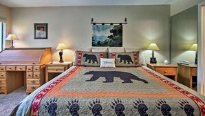 A bedroom with a bed featuring a bear-themed quilt, two bedside tables with lamps, a wooden dresser, and a framed picture on the wall.