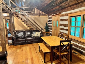 L’intérieur d’une cabane en rondins, avec un escalier en bois, un canapé en cuir, une table à manger en bois entourée de chaises et une grande fenêtre.
