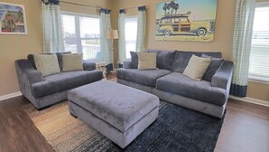 Un salon avec deux canapés gris, un pouf assorti et une grande fenêtre avec des rideaux.