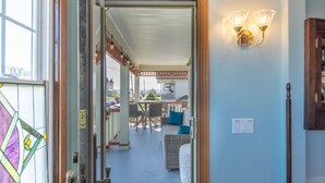Vista desde una habitación a un espacio exterior cubierto con muebles de mimbre, una mesa, una lámpara fijada a la pared y una puerta con panel de vidrio.