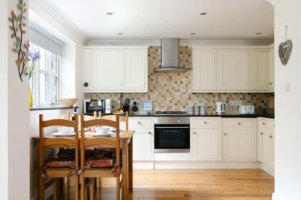 A kitchen with white cabinets, a stove, oven, and a dining area with a wooden table and chairs. - 2 Bedroom Central House Near Chapel Street (Penzance)
