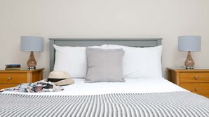 Una cama bien tendida con una colcha a rayas, una almohada gris y una almohada blanca. Hay dos mesitas de noche con lámparas y libros.