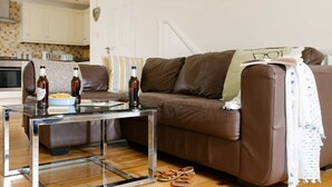 Una sala de estar acogedora con un sofá de cuero marrón, una mesa de centro de vidrio y una cocina al fondo.