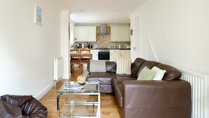Una sala de estar moderna con una mesa de centro de vidrio, sofá de cuero y vistas a una zona de cocina con armarios blancos y una estufa.