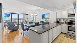A modern kitchen with white cabinets, a granite countertop, and stainless steel appliances. There is a dining area with a table and chairs, and a large window offering a view of the ocean.