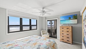 A bedroom with a bed, a chair, a dresser, a ceiling fan, a TV, and a view of the beach.