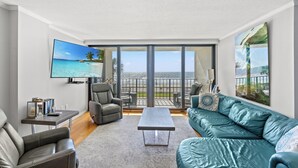 A modern living room with a large TV, comfortable seating, a coffee table, and a balcony with a view of the sea.