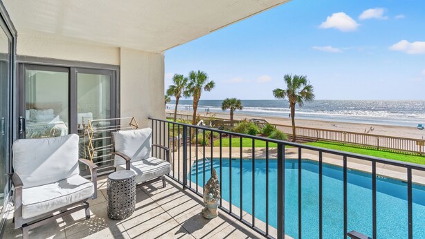 A balcony with a pool, white furniture, and a view of the beach and ocean.