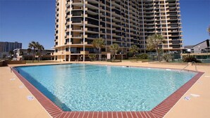 Una piscina rectangular con borde de ladrillo, rodeada por una zona arenosa. Al fondo, un edificio alto de varios pisos con balcones.