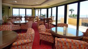 Un área de comedor con sillas y mesas estampadas, ventanas amplias con vista al mar y una alfombra roja.