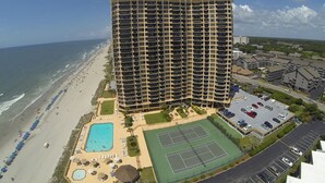 Un hotel de gran altura con varios balcones, piscina, canchas de tenis y vista al mar.