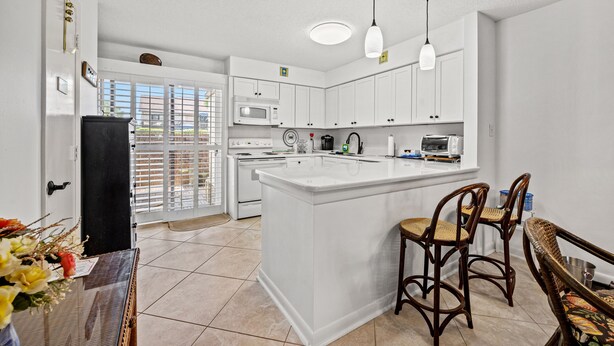 A modern kitchen with white cabinets, a central island, and a dining area with wicker chairs. - Sea Gate Villas 20 2 Bedroom Condo with Outdoor Pool! (Myrtle Beach)