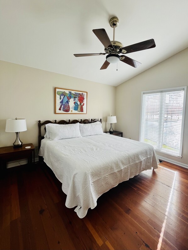 A bedroom with a wooden bed, two bedside tables with lamps, a ceiling fan, a window with blinds, and a colorful artwork on the wall.
