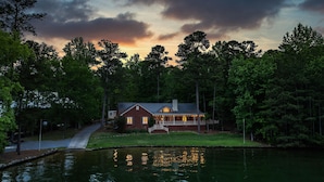 A two-story house with a porch, surrounded by trees and a body of water. - Dock. Waterslide. Beach. Firepit, Gazebo, 500ft water frontage. Views. Sleep 18. (Equality)