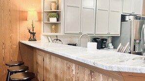 A kitchen with white cabinets, a marble countertop, a sink, a coffee maker, and a refrigerator. There are three wooden stools along the counter. - Dock. Waterslide. Beach. Firepit, Gazebo, 500ft water frontage. Views. Sleep 18. (Equality)