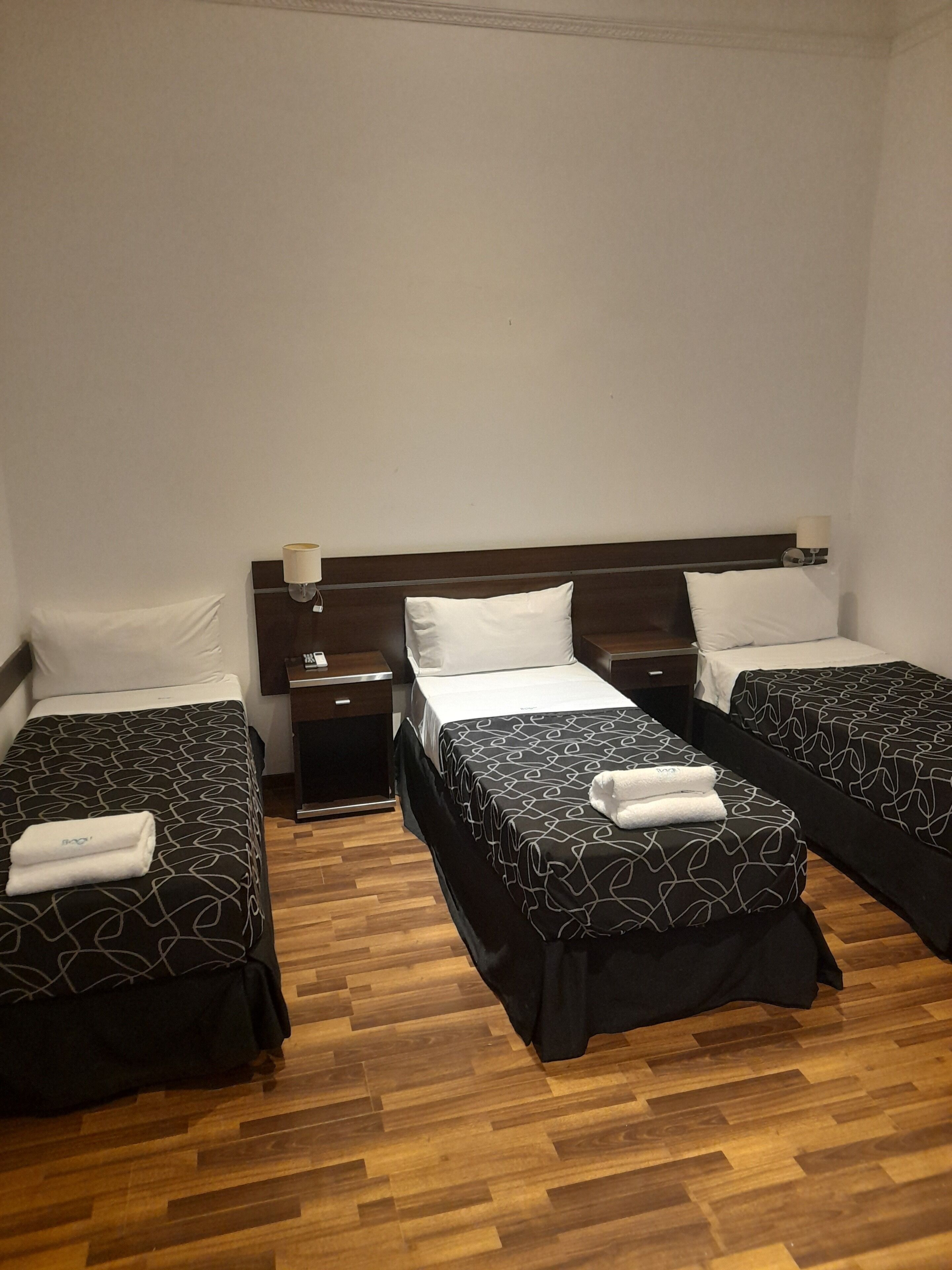 Hotellihuone, jossa on kolme erillistä sänkyä, puulattia ja yöpöytä, jossa on lamppu.
