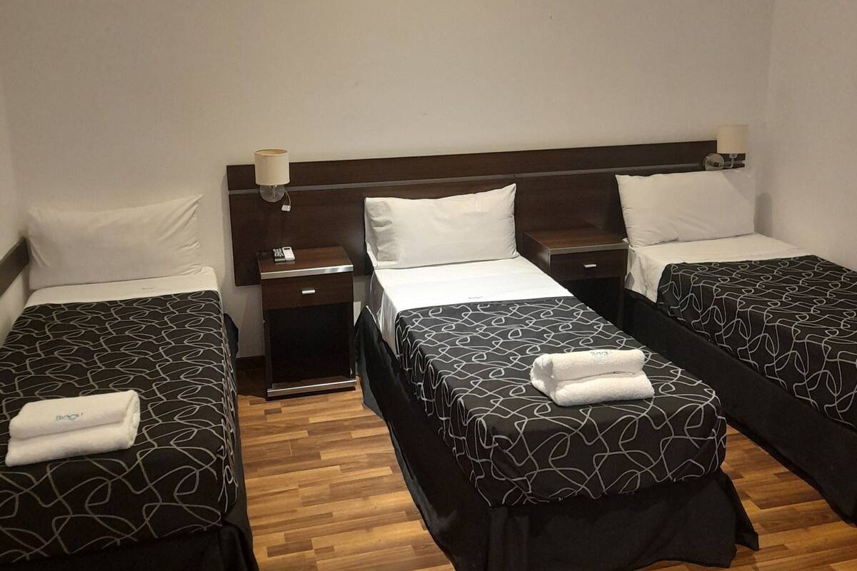Habitación de hotel con tres camas individuales, suelo de madera y una mesita de noche con lámpara.