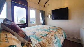 Un dormitorio con una cama cubierta por una manta estampada, un televisor de pantalla plana montado en la pared y una ventana con cortinas.