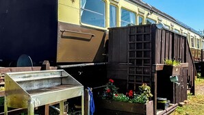 Un vagón de tren antiguo con un pequeño jardín que incluye un macetero de madera con flores y una mesa de metal.