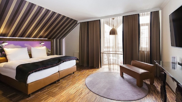 A hotel room with a bed, a desk, a chair, a TV, and a large window with curtains. - Dormero Hotel München-kirchheim Messe (Kirchheim by Munich)