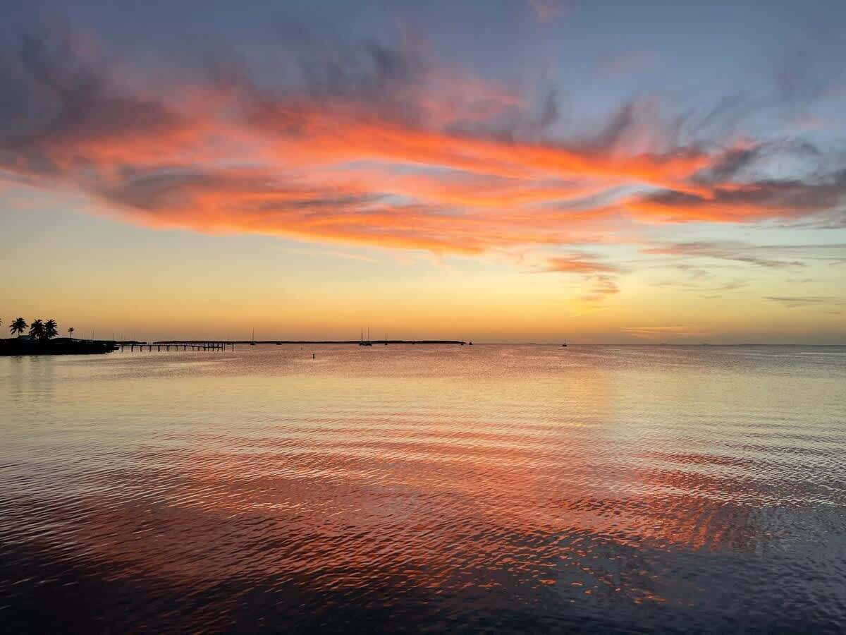 A calm body of water reflecting sunset colors, a pier extending into the water, and a distant shoreline with palm trees.