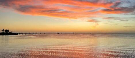A calm body of water reflecting sunset colors, a pier extending into the water, and a distant shoreline with palm trees.