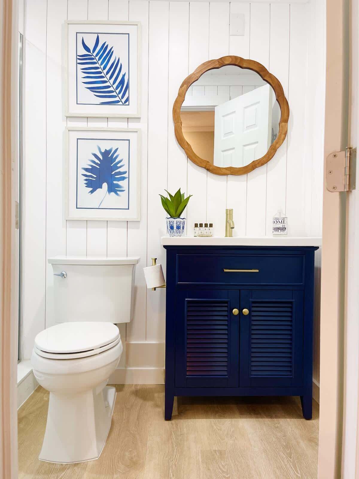 Un baño con un inodoro blanco, un tocador azul marino con un espejo redondo y obras de arte enmarcadas en la pared.