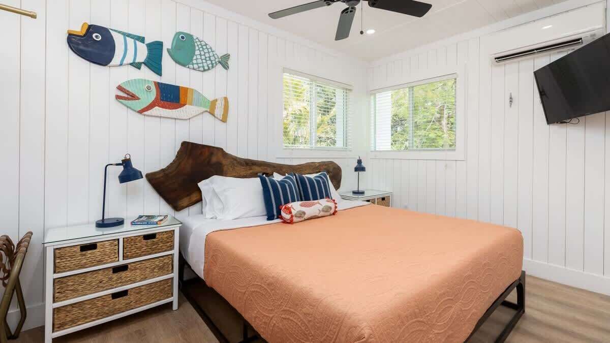 A bedroom with a wooden headboard, a bed with an orange bedspread, two nightstands with lamps, wicker storage drawers, and a wall-mounted TV.