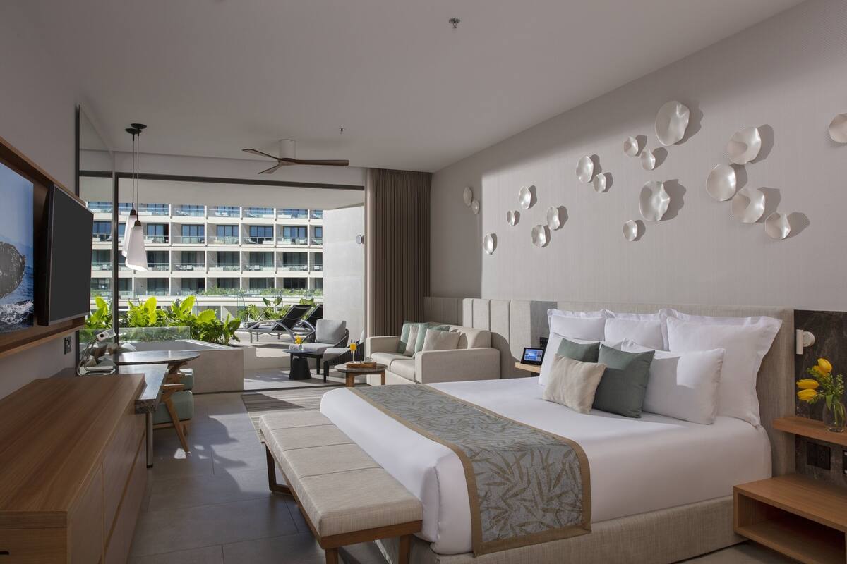 A modern hotel room with a large bed, a seating area with a table, and a balcony with plants.