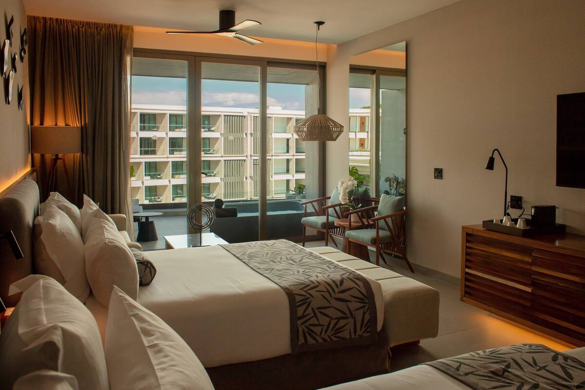 A hotel room with a large bed, a desk, a chair, and a balcony with a view of other buildings.