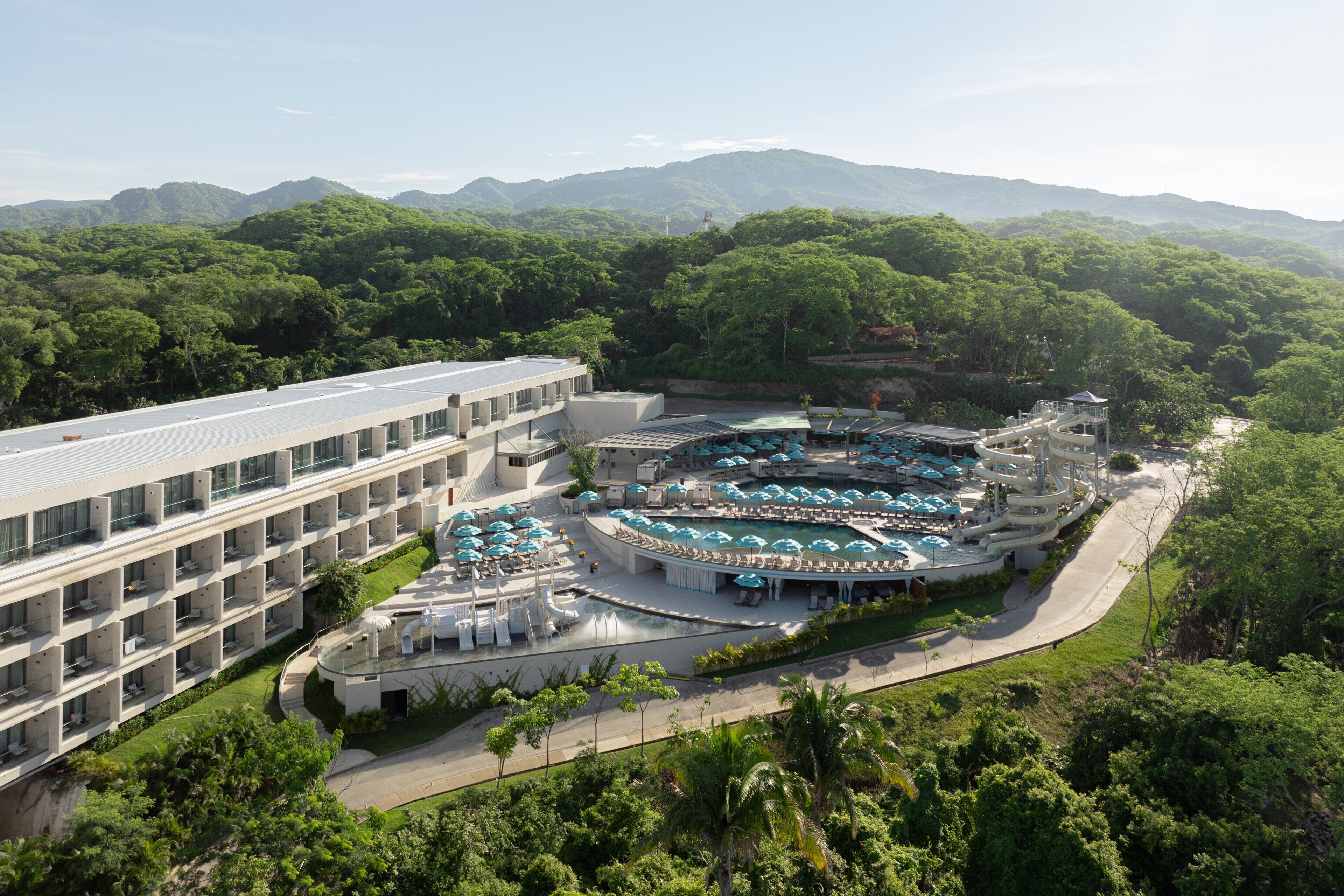 Un hotel de diseño moderno, con un amplio espacio de piscina con varios niveles y sombrillas, rodeado de exuberante vegetación y montañas al fondo.