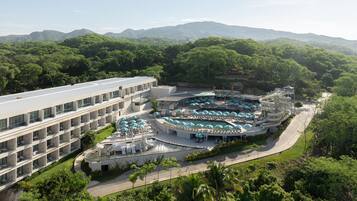 Un hotel de diseño moderno, con un amplio espacio de piscina con varios niveles y sombrillas, rodeado de exuberante vegetación y montañas al fondo.