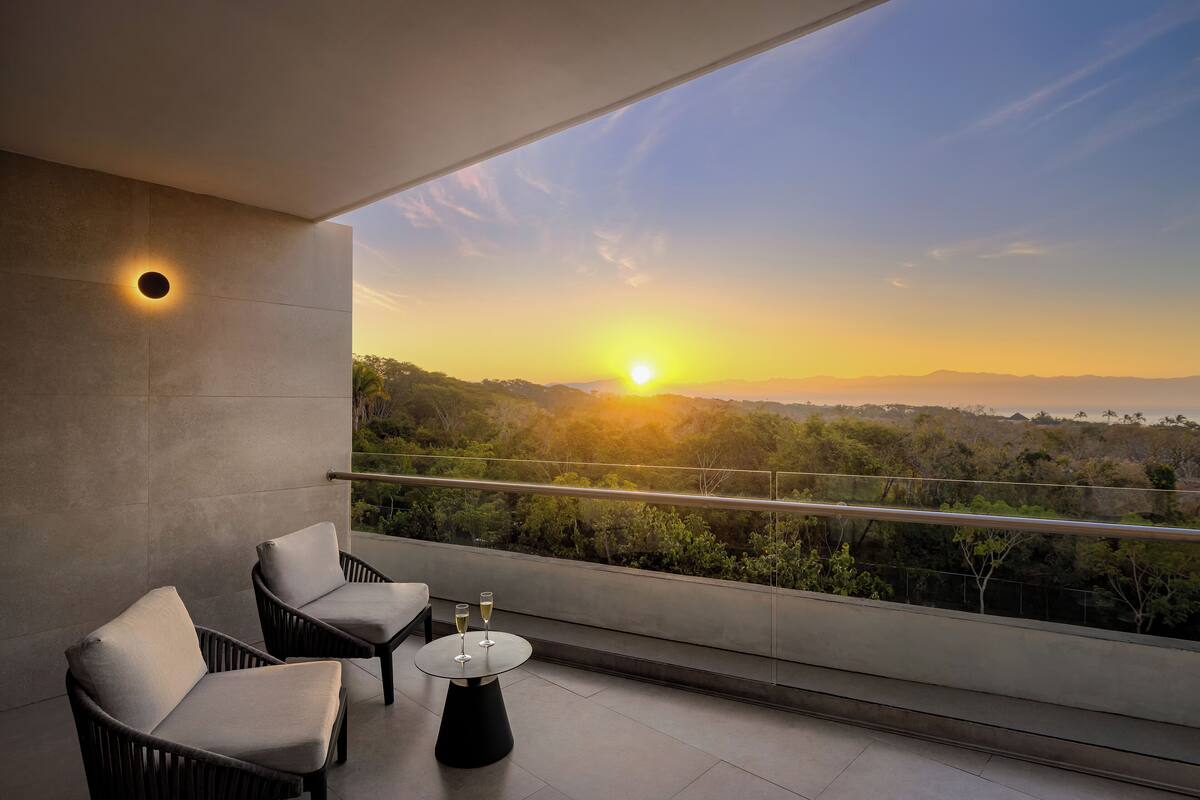 A balcony with two chairs and a table, a sunset view, and a railing.